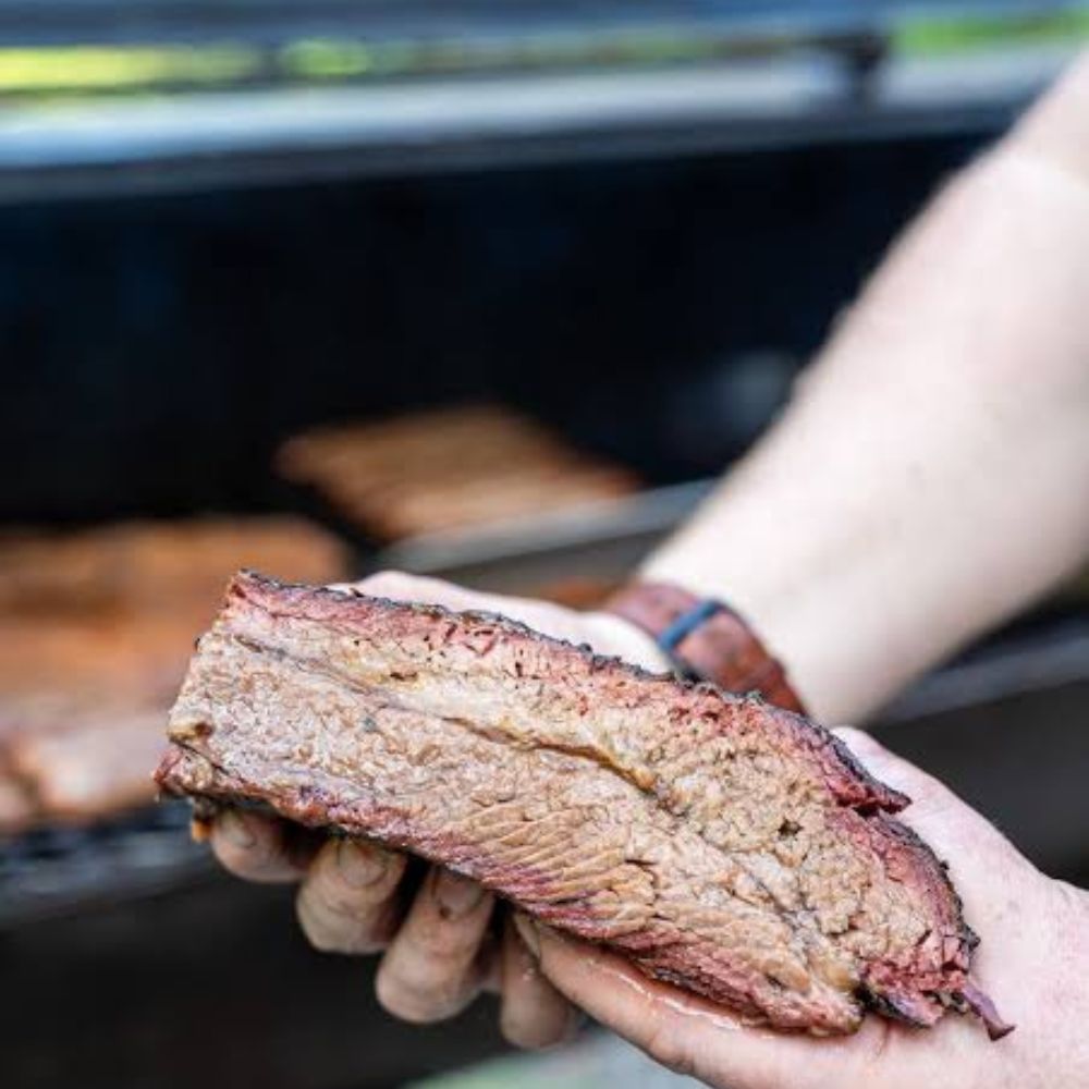 A close-up of a hand holding a well-done slice of grilled brisket in front of a grill, with another piece cooking in the background.
